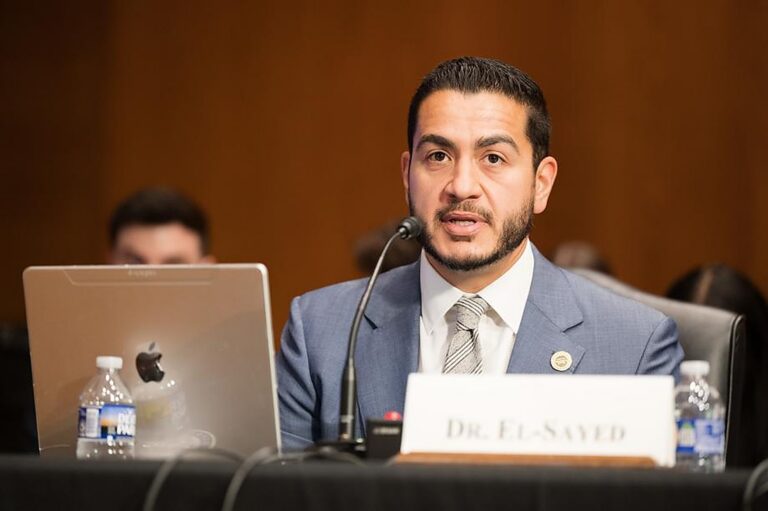 Doctor Abdul El-Sayed in a suit and tie testifying at a United States Senate hearing, speaking into a microphone behind a nameplate reading Dr. El-Sayed.