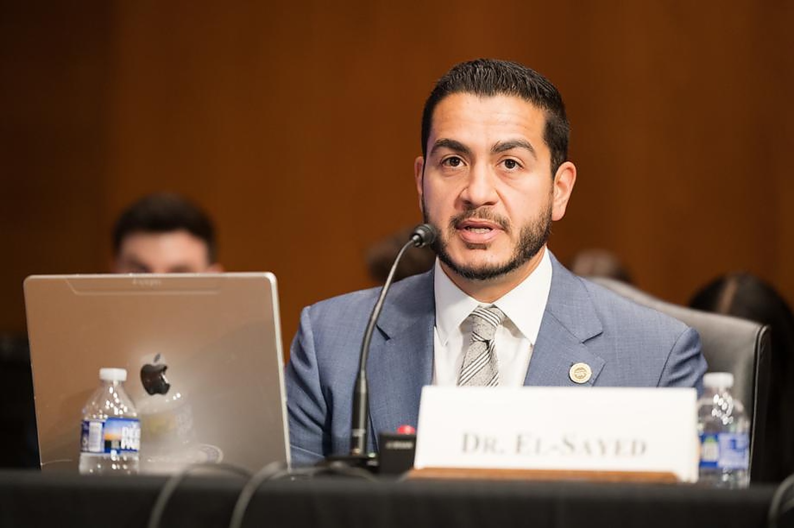 Doctor Abdul El-Sayed in a suit and tie testifying at a United States Senate hearing, speaking into a microphone behind a nameplate reading Dr. El-Sayed.